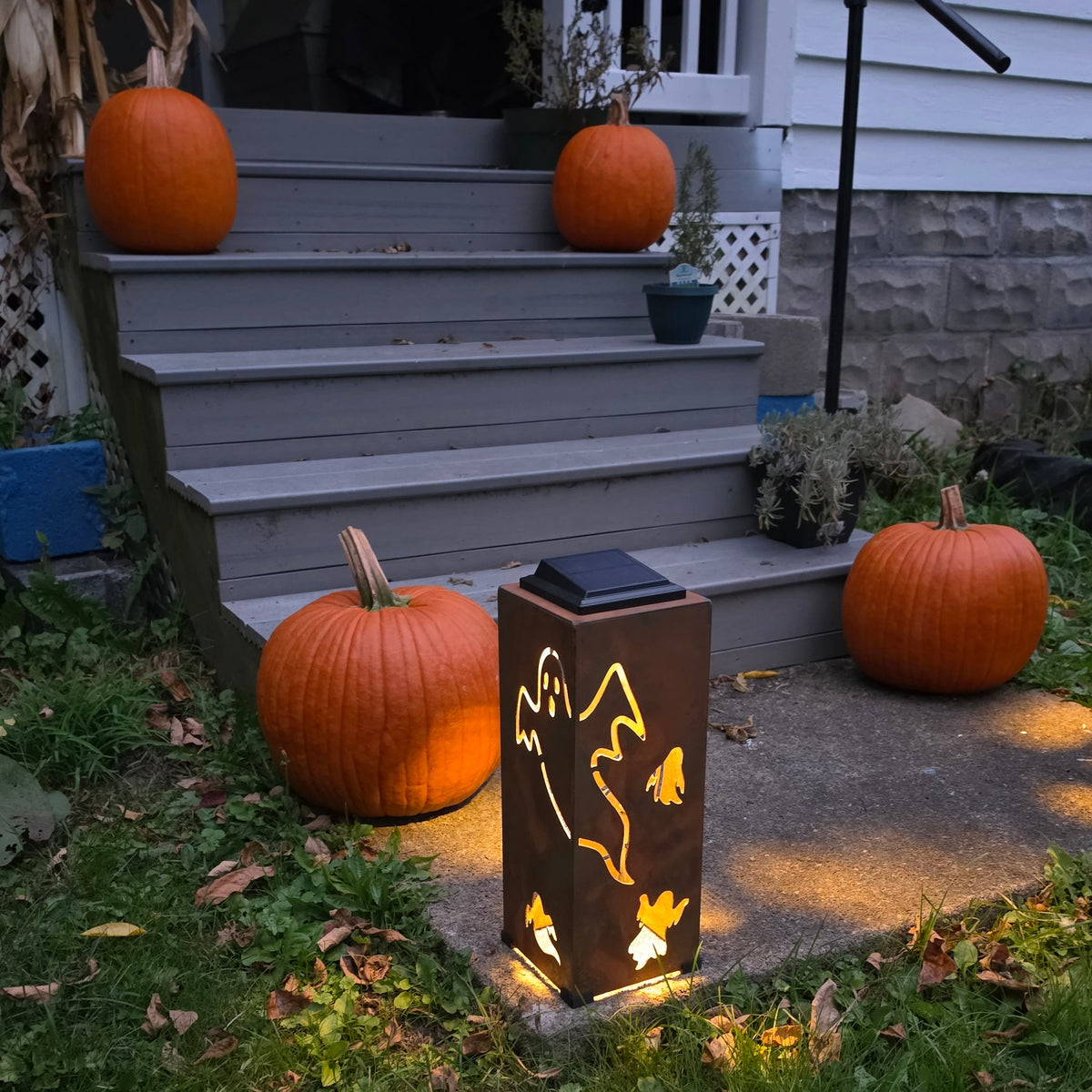 A solar-powered outdoor lantern with a mountain silhouette design stands on a concrete path in front of a house porch, casting a warm yellow glow. The scene is decorated for Halloween with several orange pumpkins on the gray steps and grassy lawn.