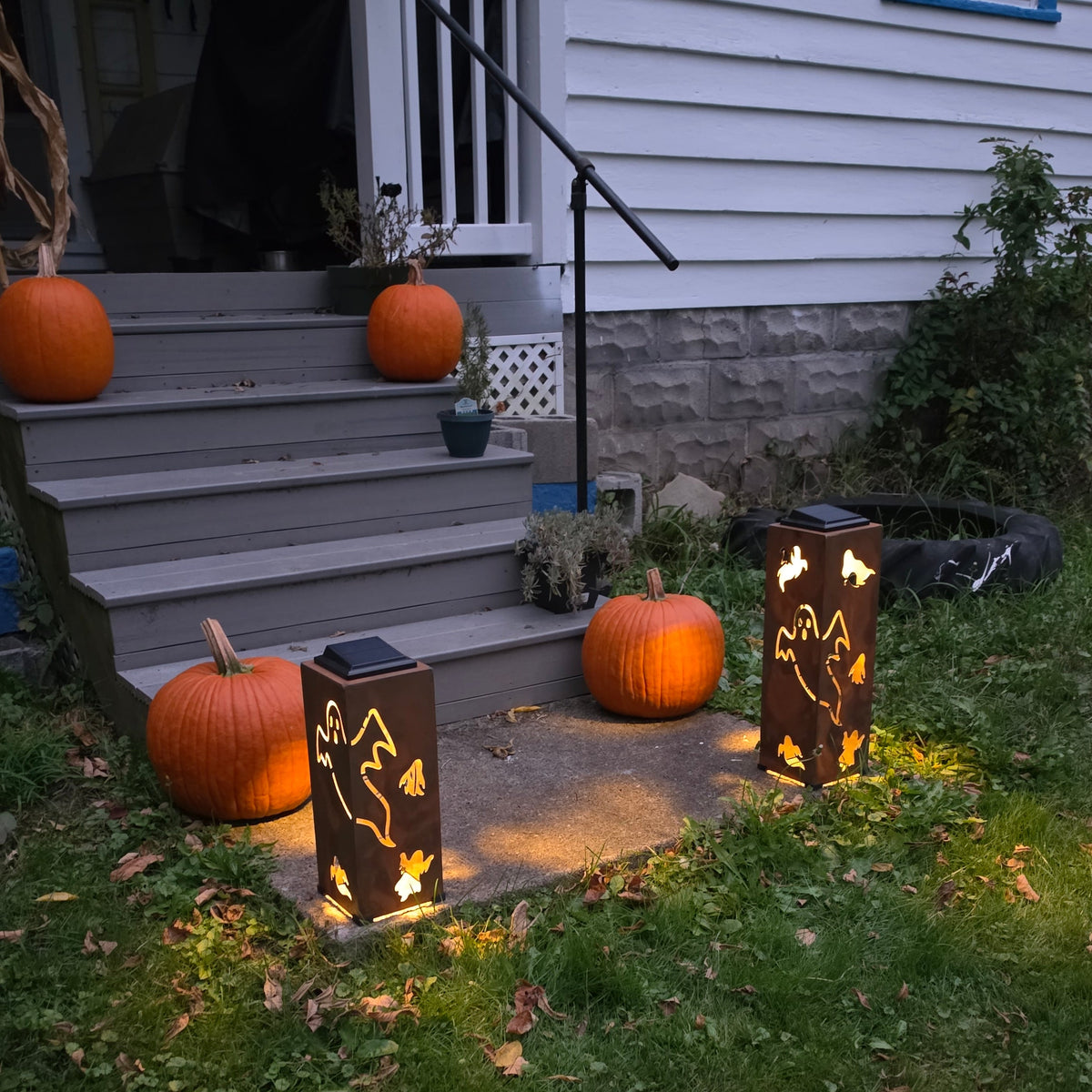 Outdoor Halloween yard scene with three decorative solar-powered lanterns placed on a front porch step and grass, each casting warm light. Pumpkins are also visible on the porch and lawn.