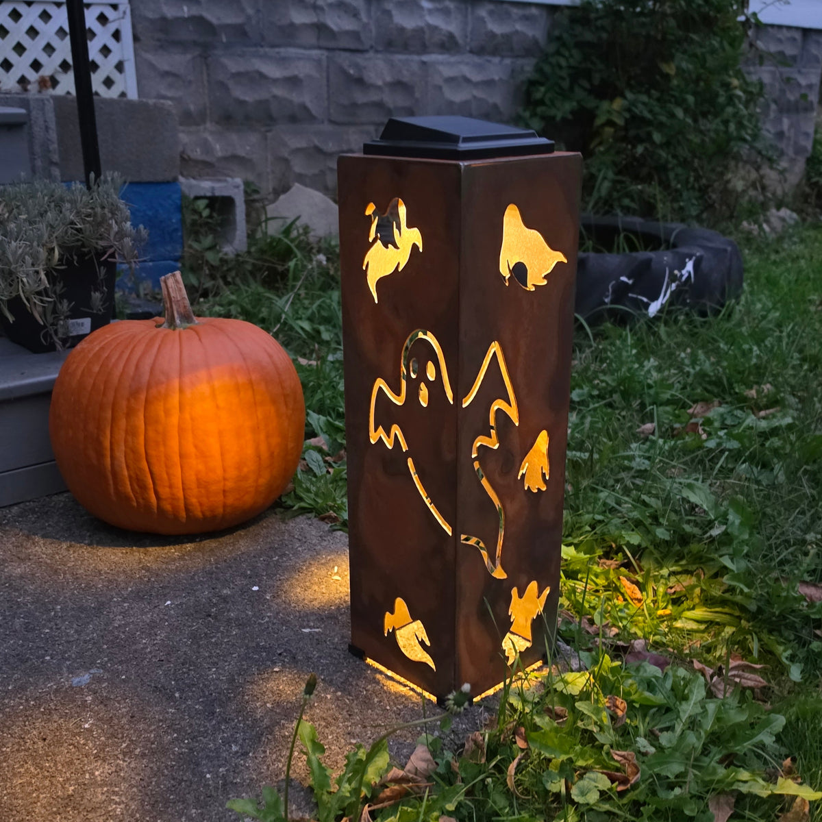 A tall rectangular outdoor light fixture with a rusted brown finish and ghost cutouts, glowing with warm light, standing in a yard next to a pumpkin and grass.