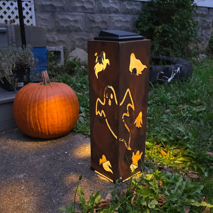 A tall rectangular outdoor light fixture with a rusted brown finish and ghost cutouts, glowing with warm light, standing in a yard next to a pumpkin and grass.