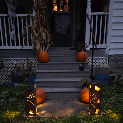 Outdoor Halloween porch scene at dusk with orange pumpkins placed on the front steps and beside two glowing black lanterns with cutout designs, set in a grassy yard in front of a house porch.