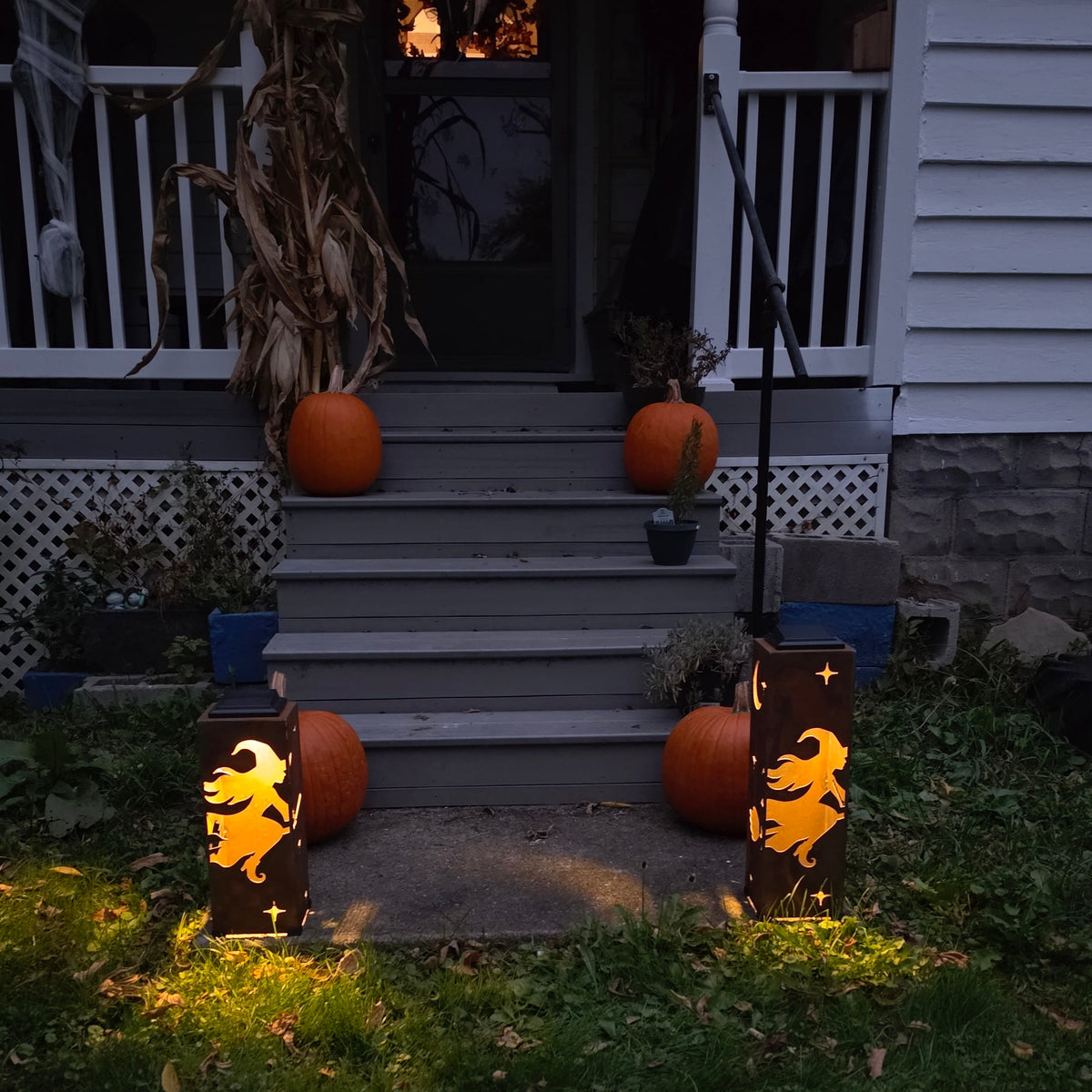 Halloween yard lights with witch silhouettes glowing orange on a grassy lawn in front of a house porch with pumpkins.