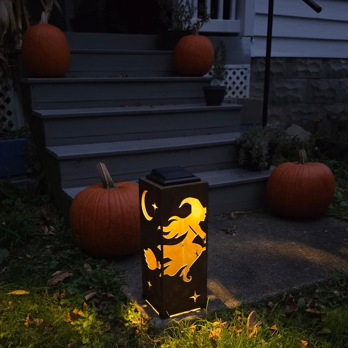 A solar-powered outdoor lantern with a witch cutting out a crescent moon and star pattern, glowing with warm yellow light on a grassy lawn in front of a house porch decorated with pumpkins.