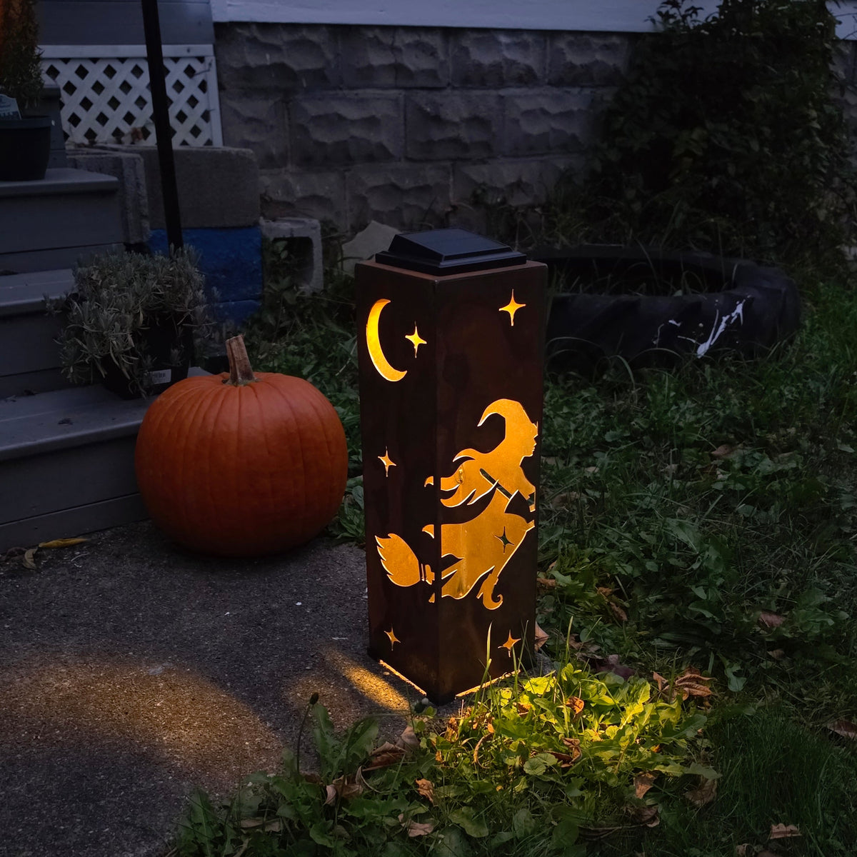 A solar-powered outdoor ground light with a Halloween design of a witch riding a broomstick under a crescent moon and stars, casting a warm yellow glow on grass and pavement next to a pumpkin in a yard.