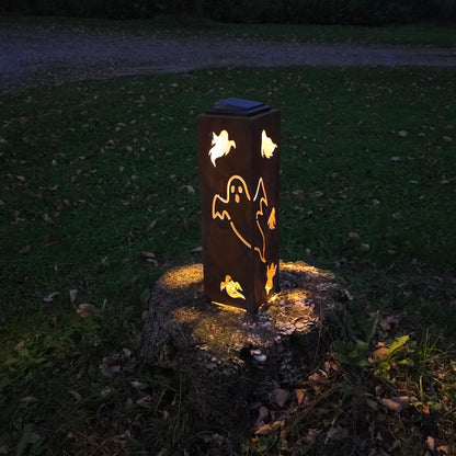 A solar-powered outdoor light fixture with a ghost cutout design, glowing warmly on a tree stump in a grassy yard at dusk.
