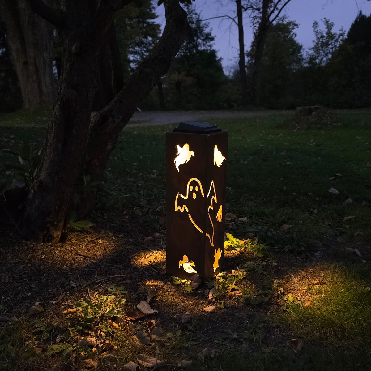 A dark metal outdoor stake light with ghost cutouts, glowing warmly at dusk in a grassy yard near a tree.