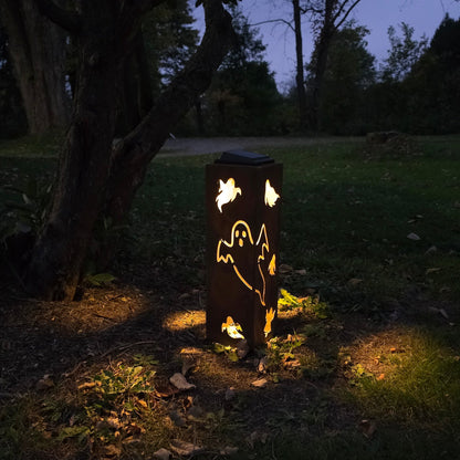 A dark metal outdoor stake light with ghost cutouts, glowing warmly at dusk in a grassy yard near a tree.