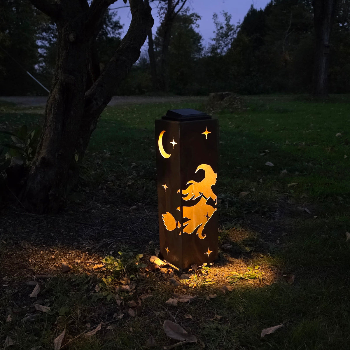 A dark outdoor lantern with a witch riding a broomstick and a crescent moon design, glowing warmly on a grassy lawn at night under trees.