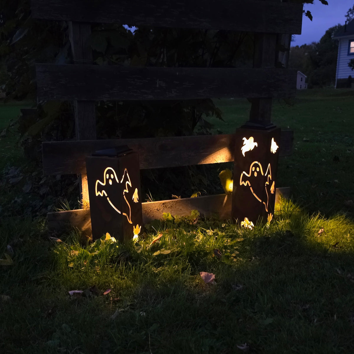 Two glowing outdoor lanterns with ghost cutout designs, placed on grass in front of a wooden fence at dusk.