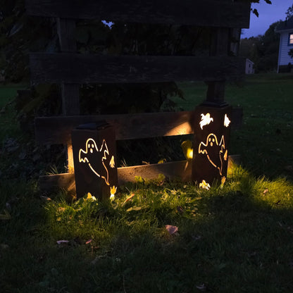 Two glowing outdoor lanterns with ghost cutout designs, placed on grass in front of a wooden fence at dusk.