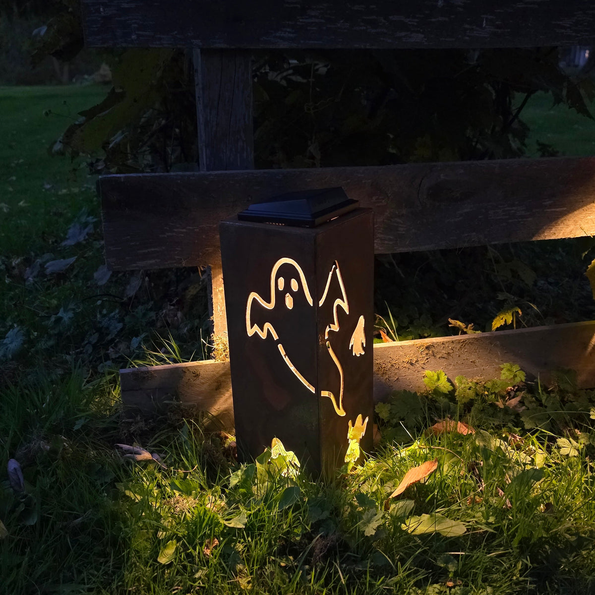 A dark metal outdoor lantern with a ghost cutout, glowing warmly on a grassy lawn next to a wooden fence at night.