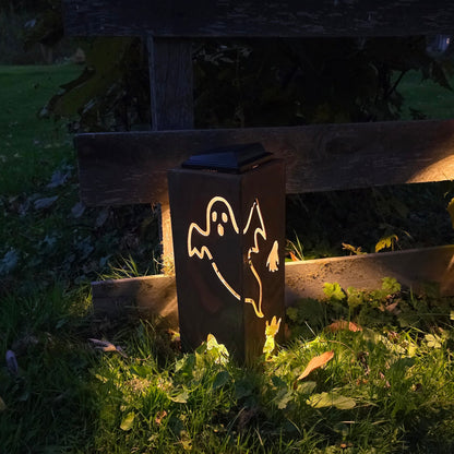 A dark metal outdoor lantern with a ghost cutout, glowing warmly on a grassy lawn next to a wooden fence at night.