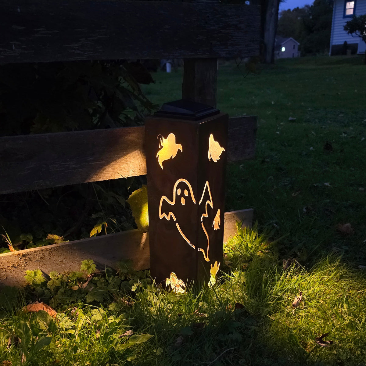A dark outdoor lantern with butterfly and butterfly-like cutout designs, illuminated at dusk on a grassy lawn beside a wooden fence.
