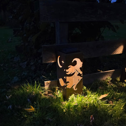 A solar-powered outdoor ground lantern with a cutout design, emitting a warm glow on the grass in a dark garden setting.
