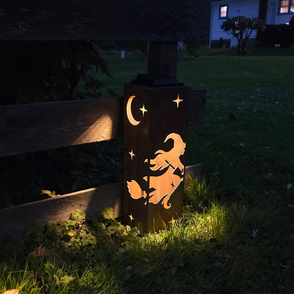 A decorative outdoor light post at dusk, featuring a cutout design of a witch riding a broomstick under a crescent moon and stars, casting a warm glow on the grass beside a wooden fence.