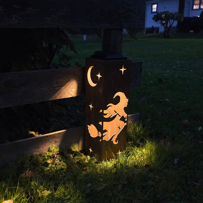 A decorative outdoor light post at dusk, featuring a cutout design of a witch riding a broomstick under a crescent moon and stars, casting a warm glow on the grass beside a wooden fence.