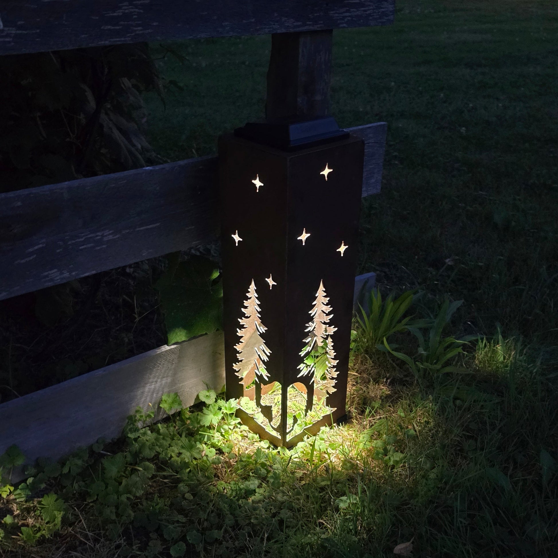 Outdoor solar light with a dark metal silhouette of two pine trees and stars, casting a warm yellow glow on grass at night next to a wooden fence.