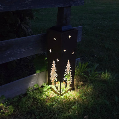 Outdoor solar light with a dark metal silhouette of two pine trees and stars, casting a warm yellow glow on grass at night next to a wooden fence.