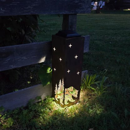 A rustic metal outdoor garden light shaped like a lantern with star cutouts and a tree silhouette, glowing on a grassy lawn at dusk next to a wooden fence.