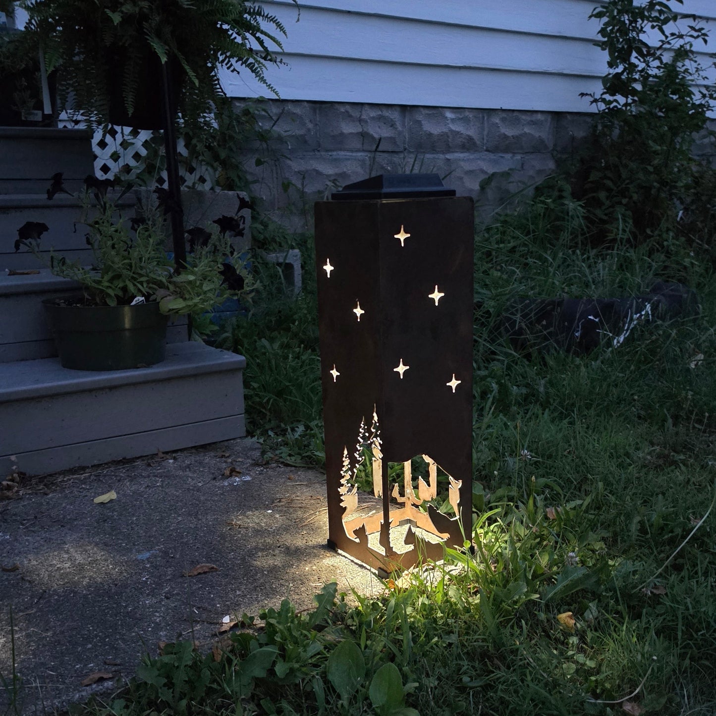 A dark metal outdoor lantern with star and tree cutouts, illuminated from within, standing on grass beside a house exterior at night.