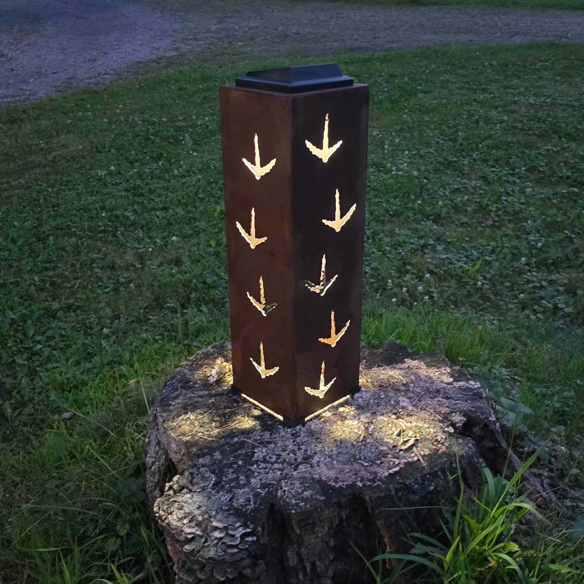 A rectangular outdoor solar light with a dark metal body and glowing warm turkey print cutouts, displayed on a tree stump in a grassy backyard at dusk.