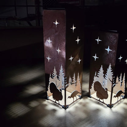 Two tall rectangular metal lanterns with laser-cut silhouettes of pine trees, turkeys, and stars, glowing with warm light in a dark indoor setting.