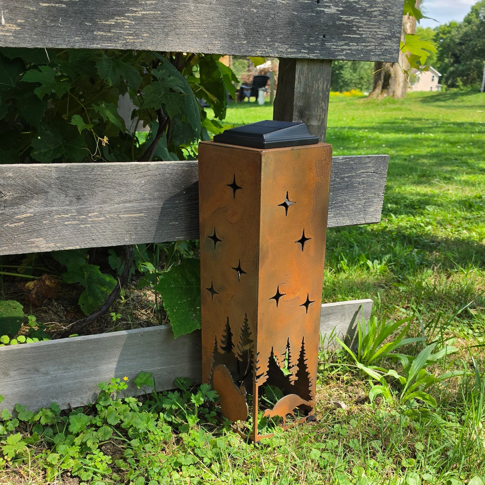 A rusted metal outdoor light fixture with star and tree cutouts, placed on grass next to a wooden fence in a yard.
