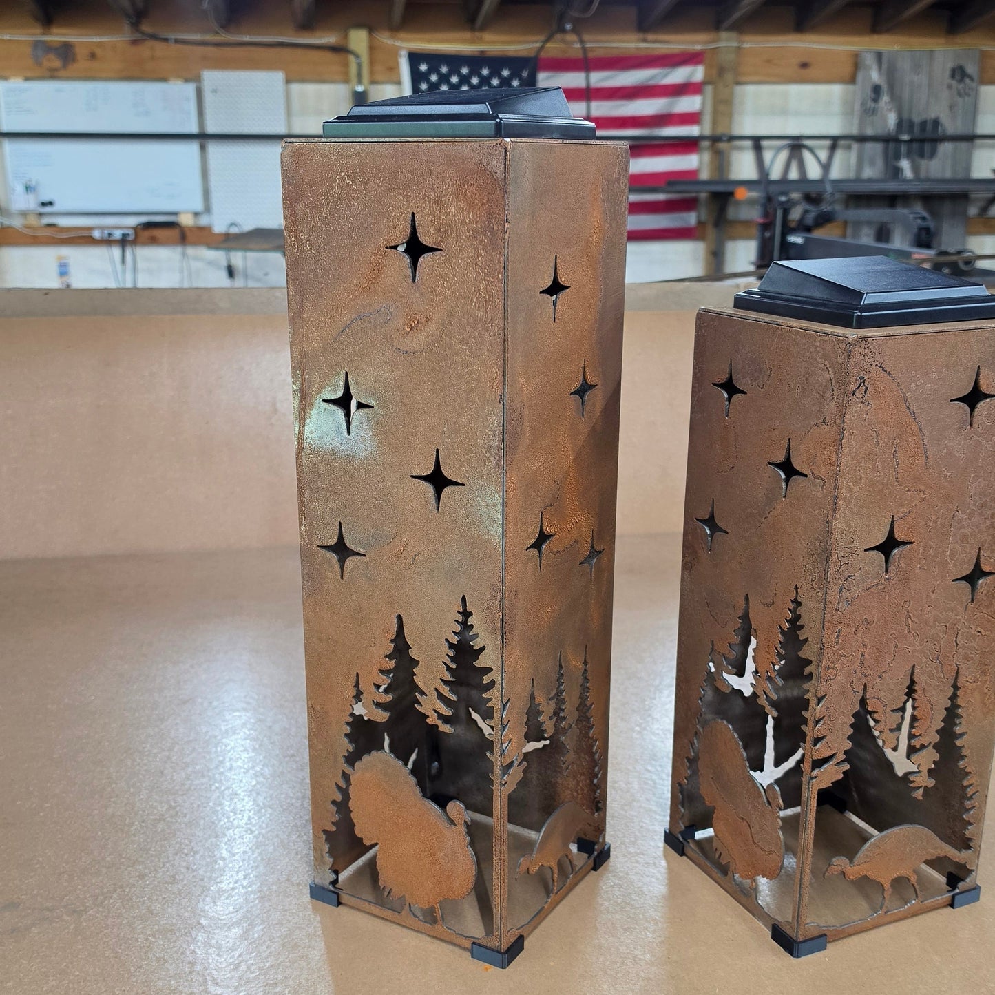 Two rustic metal lanterns with laser-cut forest and turkey silhouettes, displayed on a beige floor inside a workshop.