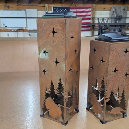Two rustic metal lanterns with laser-cut forest and turkey silhouettes, displayed on a beige floor inside a workshop.