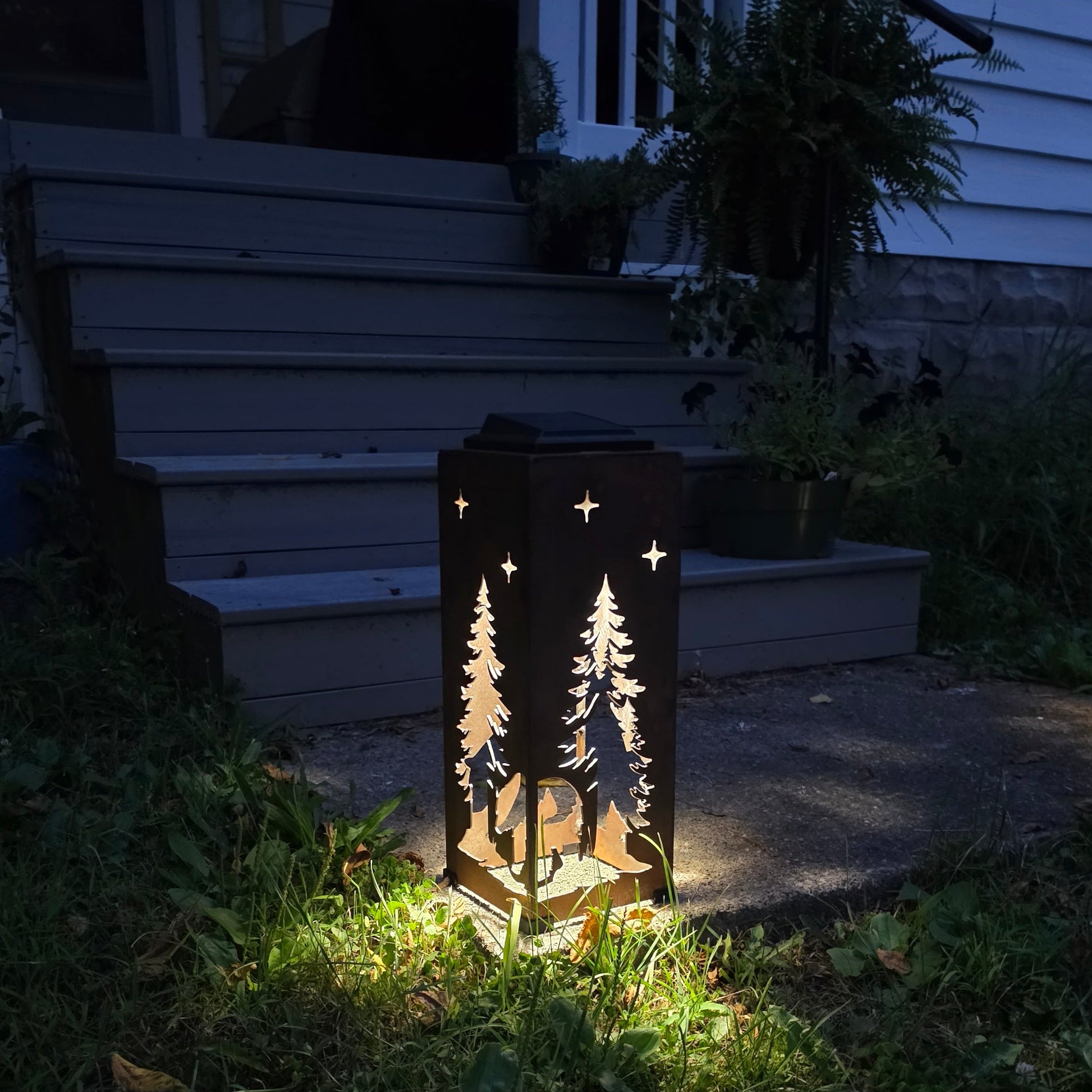 A rustic metal outdoor lantern with laser-cut tree and star designs, glowing with warm light on a grassy path in front of a house porch at night.