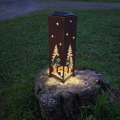 A rectangular outdoor lantern with a dark brown rust finish, featuring laser-cut pine tree and star designs, glowing warmly from within. It is shown lit up on a tree stump in a grassy backyard at dusk.