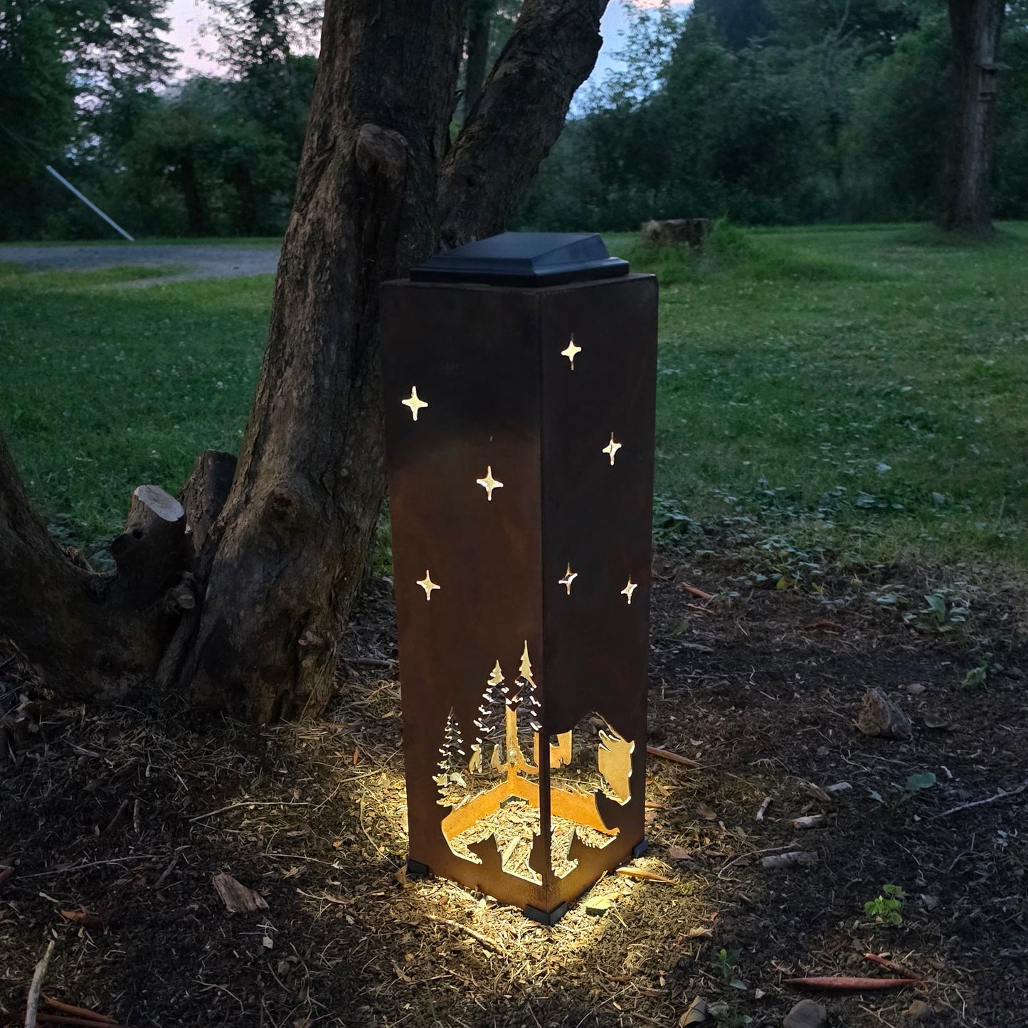 A tall rectangular outdoor lantern with a dark metal finish stands on the ground beside a tree in a grassy yard at dusk. The lantern is lit from within, casting a warm glow, and has star-shaped cutouts on the sides plus a forest silhouette near the bottom.