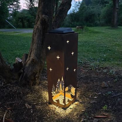 A tall rectangular outdoor lantern with a dark metal finish stands on the ground beside a tree in a grassy yard at dusk. The lantern is lit from within, casting a warm glow, and has star-shaped cutouts on the sides plus a forest silhouette near the bottom.