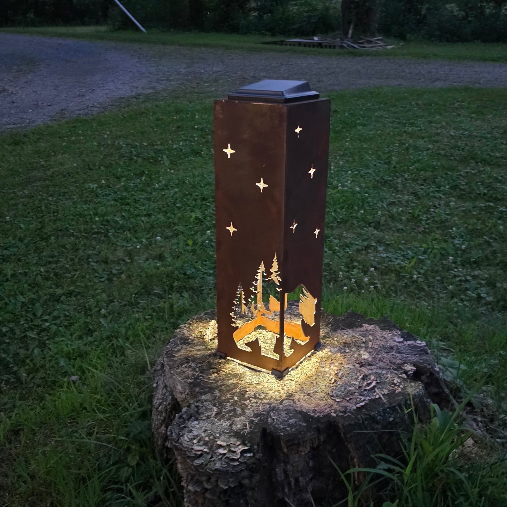 A rectangular metal outdoor lantern with star and tree cutouts, glowing with warm light from within, placed on a tree stump in a grassy yard at dusk.
