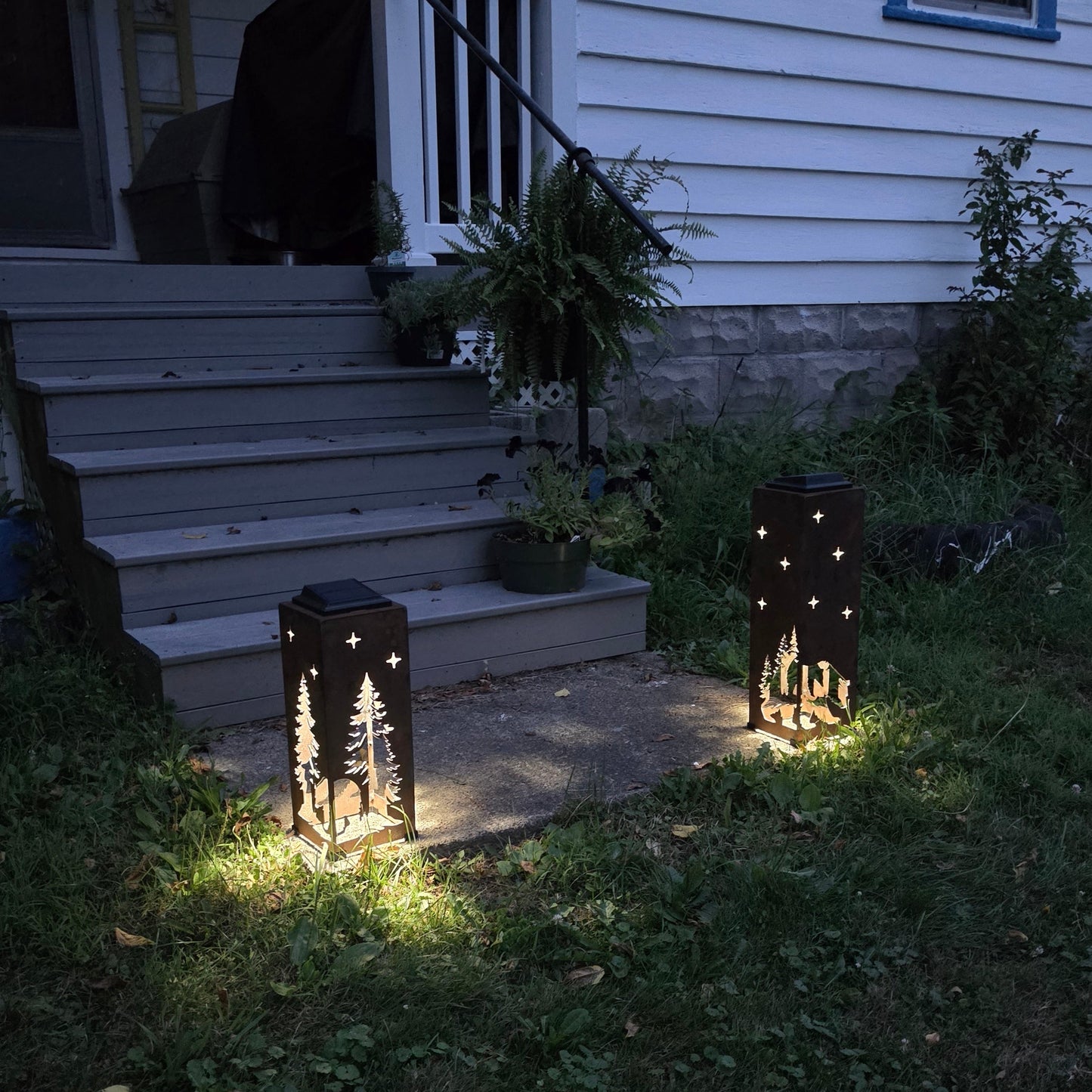 Two rustic outdoor lanterns with star and tree cutout designs are placed on a grassy yard path at night, casting warm light on the ground. The scene is set in front of a house with gray steps and white siding.