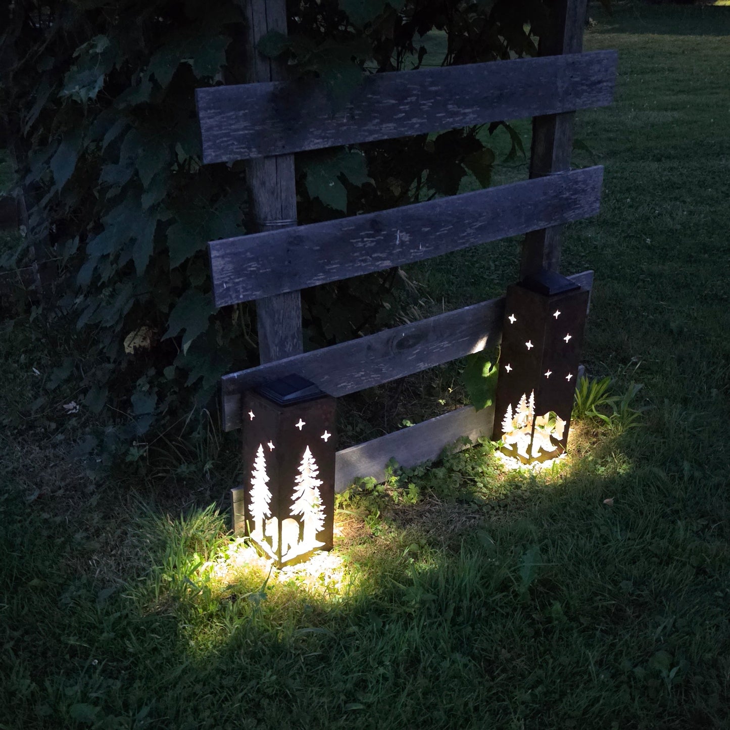 Two decorative outdoor lanterns with star cutouts are placed on a grassy lawn at night, casting warm light on the ground. One lantern features a tree silhouette design, and both are set against a rustic wooden fence backdrop.