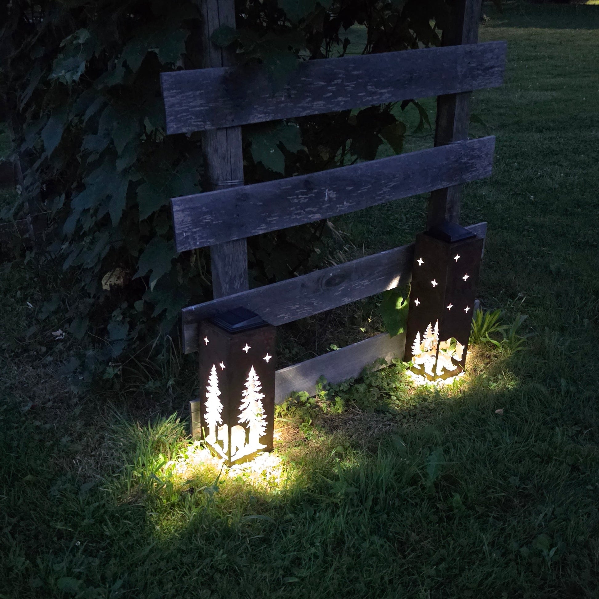 Two decorative outdoor lanterns with star cutouts are placed on a grassy lawn at night, casting warm light on the ground. One lantern features a tree silhouette design, and both are set against a rustic wooden fence backdrop.