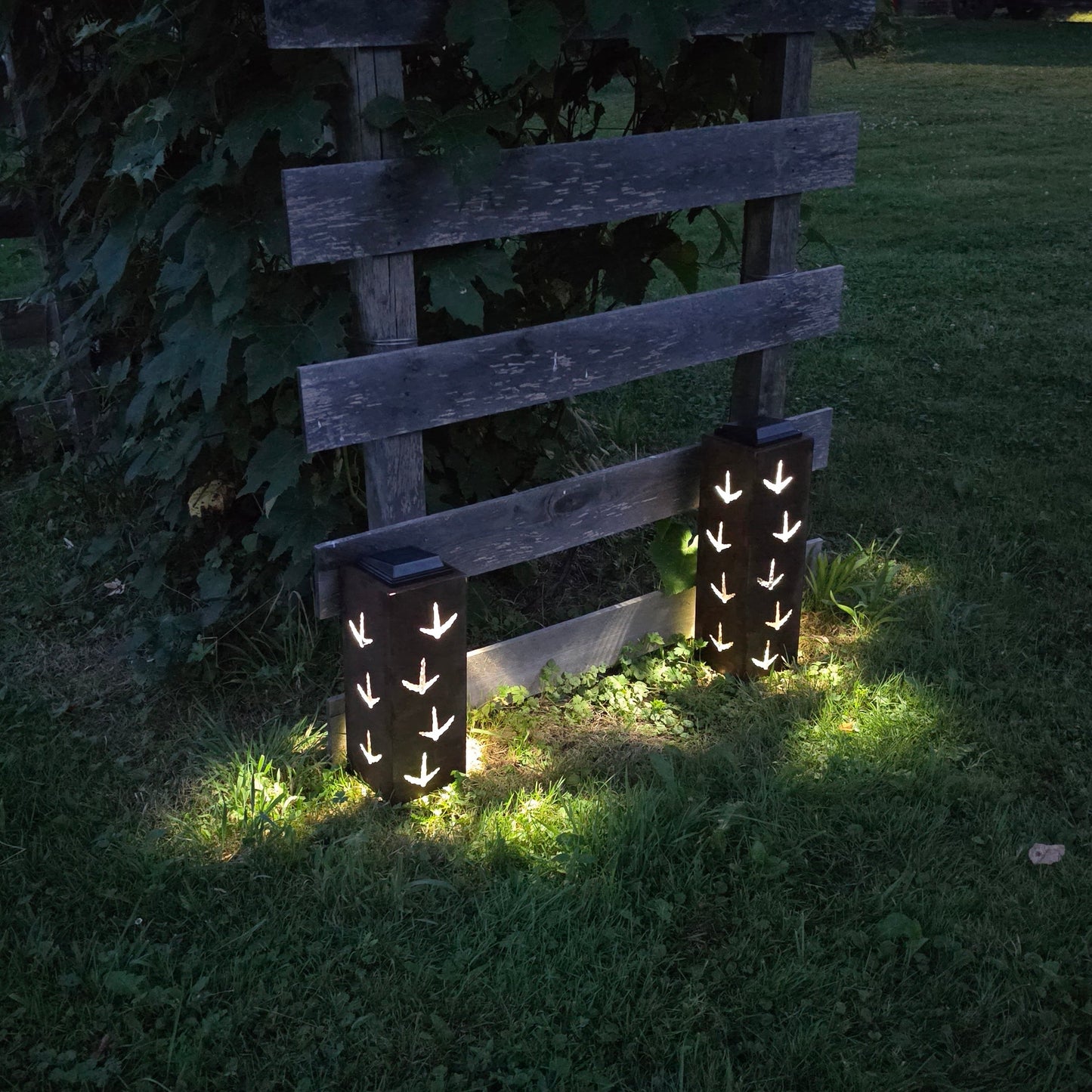 Rustic metal solar pillars with turkey prints on them glowing at dusk in front of a grape vine