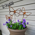 Three rusted metal butterfly decorative elements with a pot of purple flowers against a wooden background.
