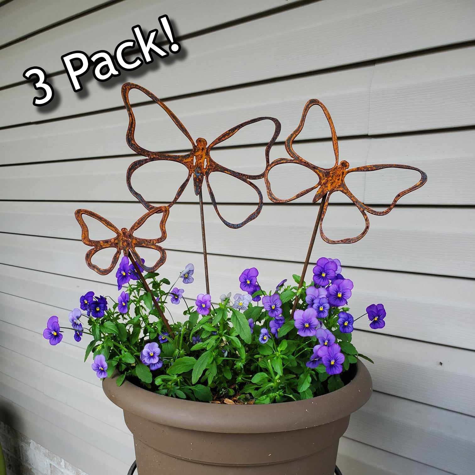 Three rusted metal butterfly decorative elements with a pot of purple flowers against a wooden background.