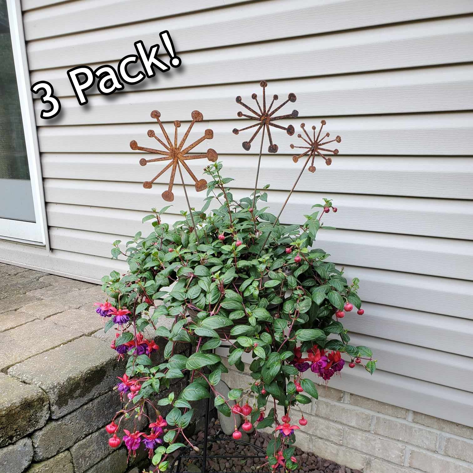 Three rust-colored metal starburst garden stakes in front of a potted plant with flowers against a beige wall.