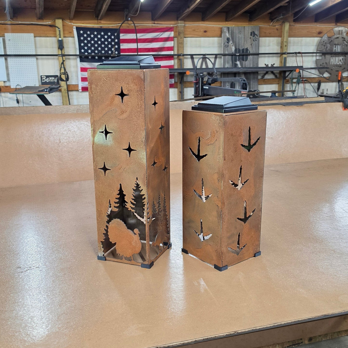 Two rustic metal lanterns with laser-cut pine tree and turkey designs, displayed on a wooden workbench inside a workshop with an American flag in the background.