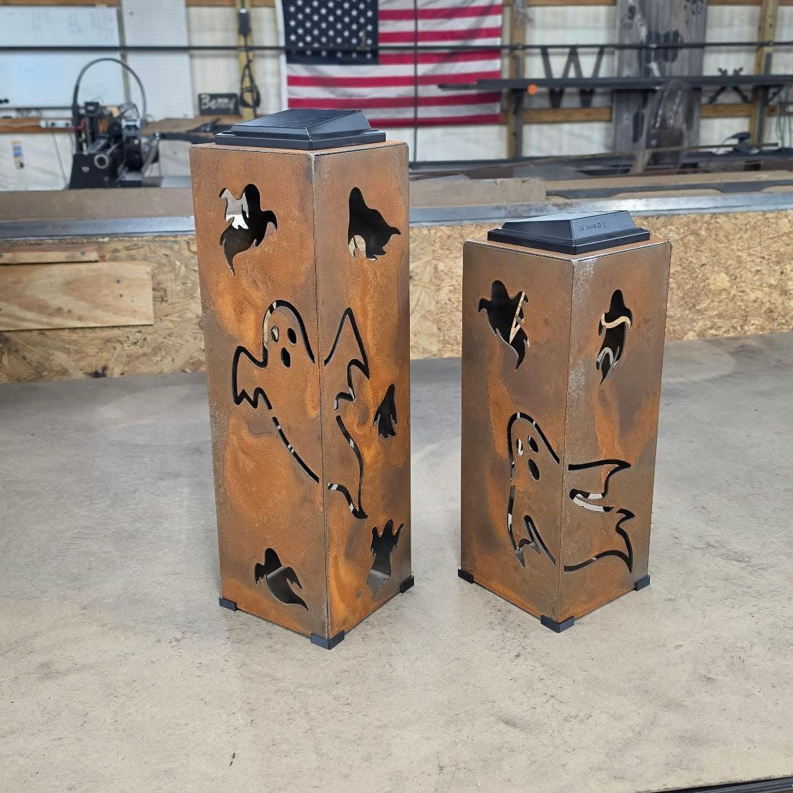 Two rustic metal ghost-shaped lanterns with black cutout details, standing on a workshop floor in front of a wooden ceiling and an American flag.