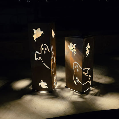 Two tall rectangular metal lanterns with ghost cutouts, casting warm light and shadow on a dark surface in a dim setting.
