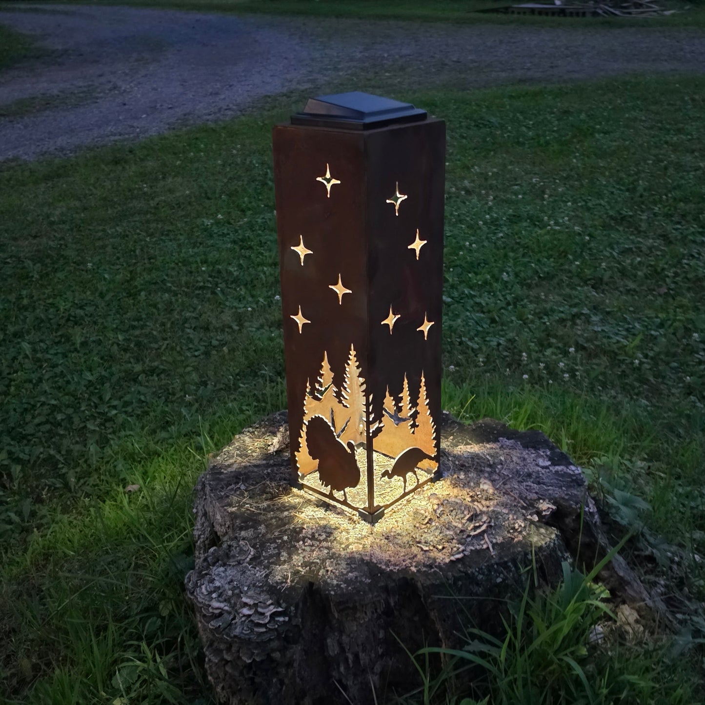 A solar-powered outdoor lantern with a laser-cut design of trees, turkeys, and stars, glowing warmly on a tree stump in a grassy yard at dusk.