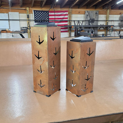 Two rustic metal display pedestals with cutout turkey print patterns, standing on a wooden platform in a workshop with an American flag in the background.
