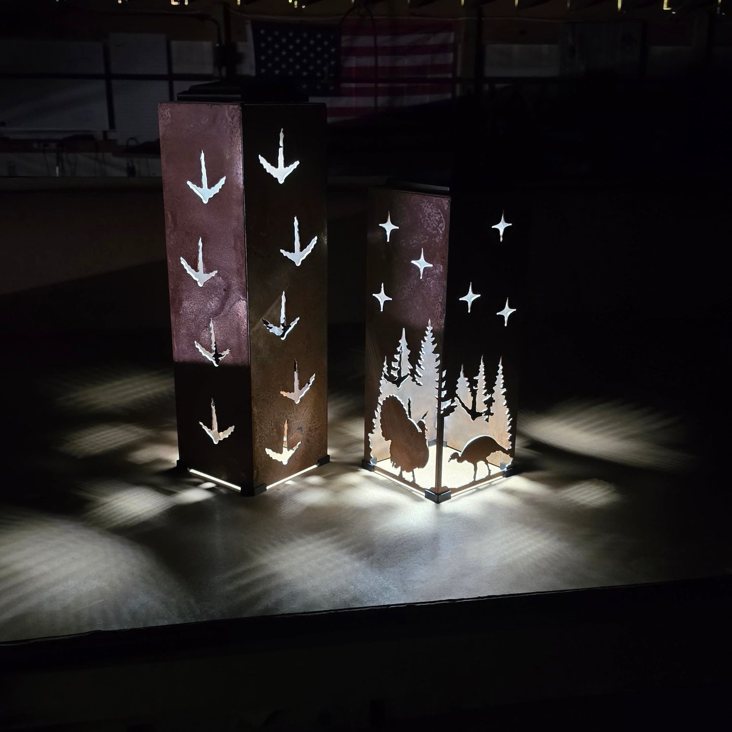 Two tall rectangular decorative lamps or panels standing on a dark stage, illuminated to show white bird and tree silhouettes against a black background.