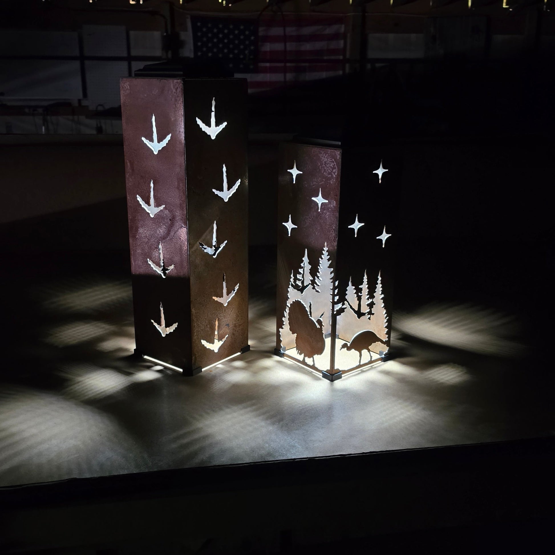 Two tall rectangular decorative lamps or panels standing on a dark stage, illuminated to show white bird and tree silhouettes against a black background.