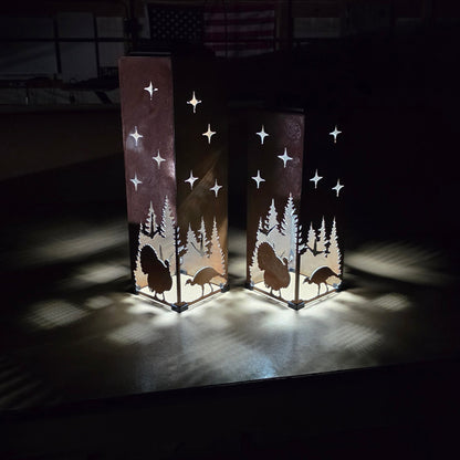 Two tall rectangular rustic lanterns with laser-cut silhouettes of trees, turkeys, and stars, glowing with warm light in a dark indoor setting.