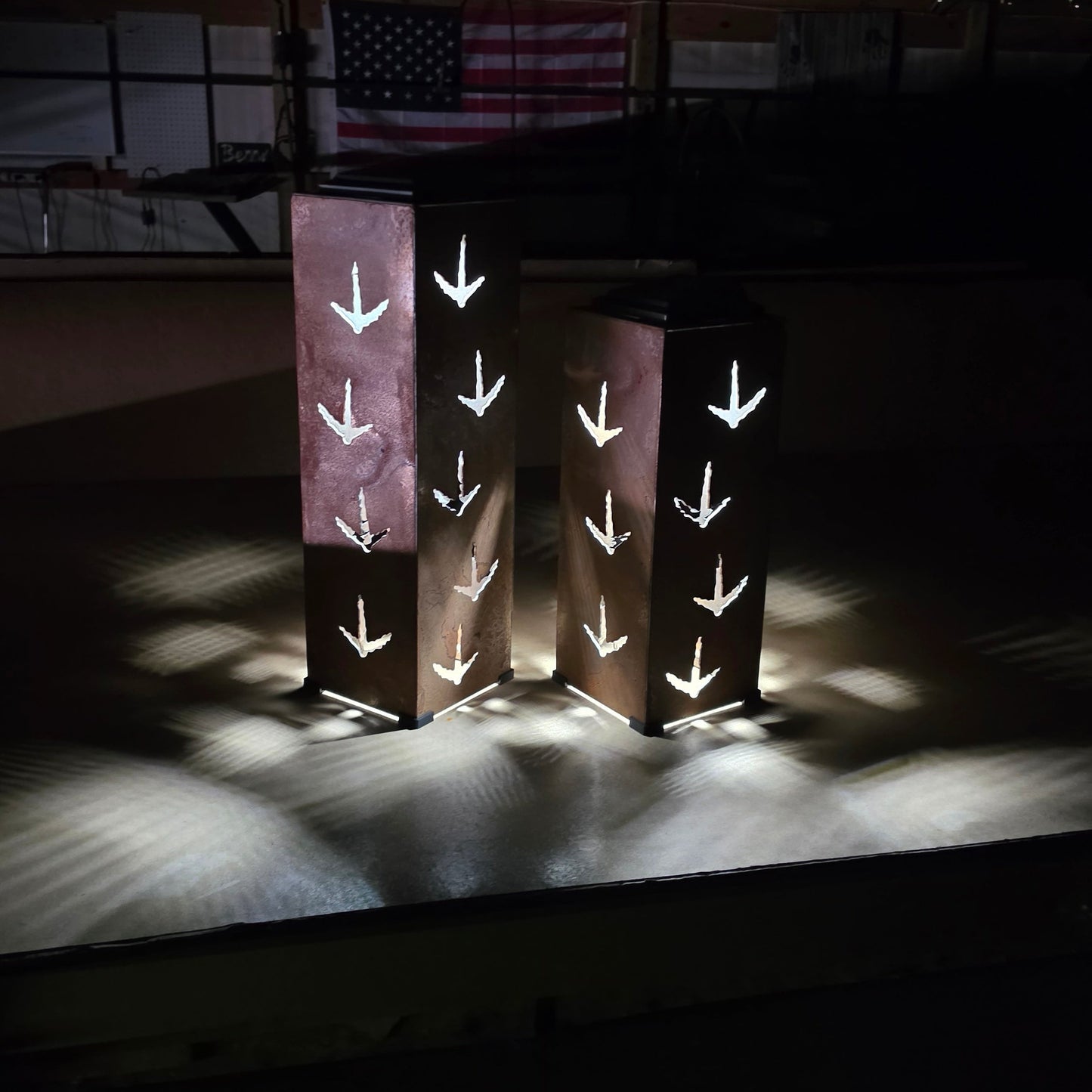 Two tall rectangular metal lanterns with cutout turkey print patterns, glowing with warm light on a dark stage-like setting with an American flag in the background.